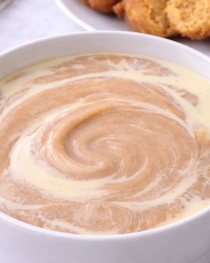 A bowl of smooth pap swirled with milk, with akara in the background